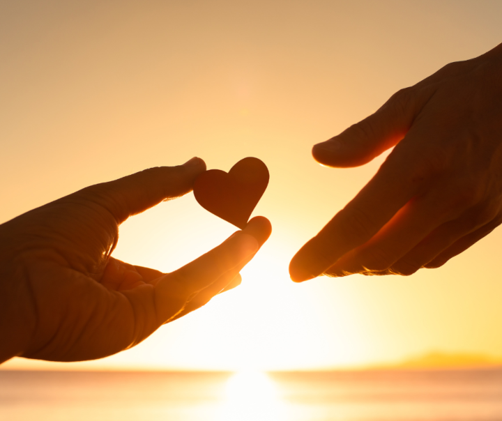 One hand handing a heart-shaped object out to another hand, with the bright sun in the background.